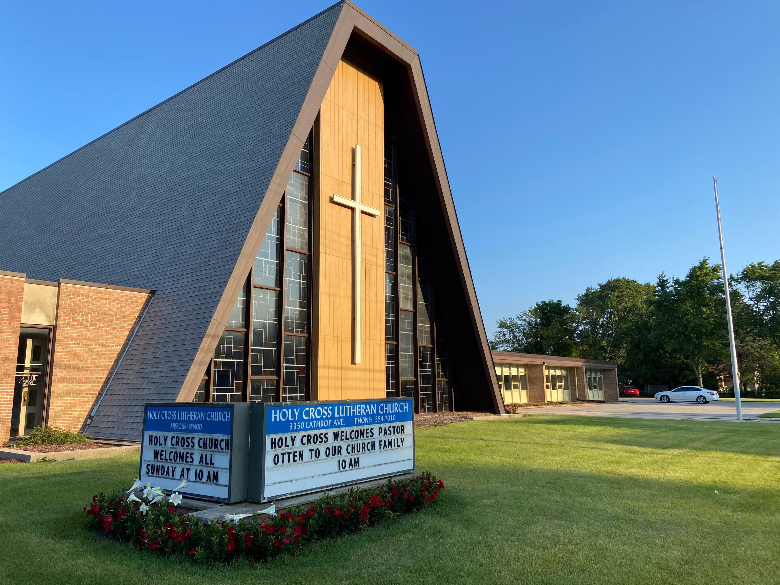 An exterior shot of Holy Cross Lutheran Church in Racine, Wisconsin. They have an A-frame roof, with stained glass on the left and right sides of the front, flanking a large cross in the middle. They have a sign out front welcoming Pastor Otten and his fam