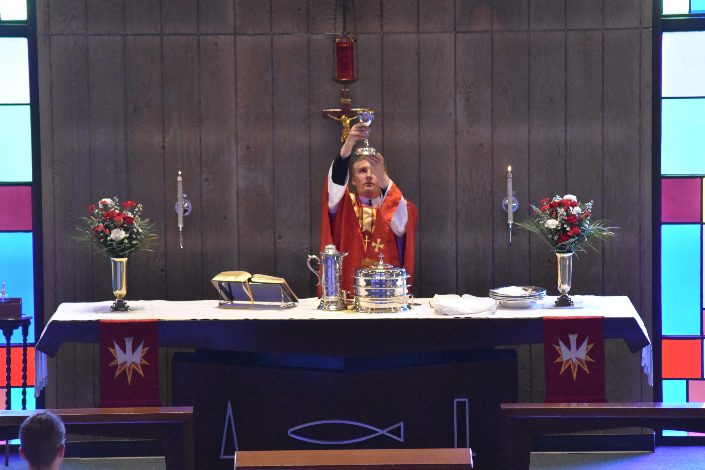 Pastor Luke Otten elevates the chalice in a service at Holy Cross Lutheran Church in Racine, Wisconsin. He is wearing a red chasuble. The altar also has red paraments and flowers.