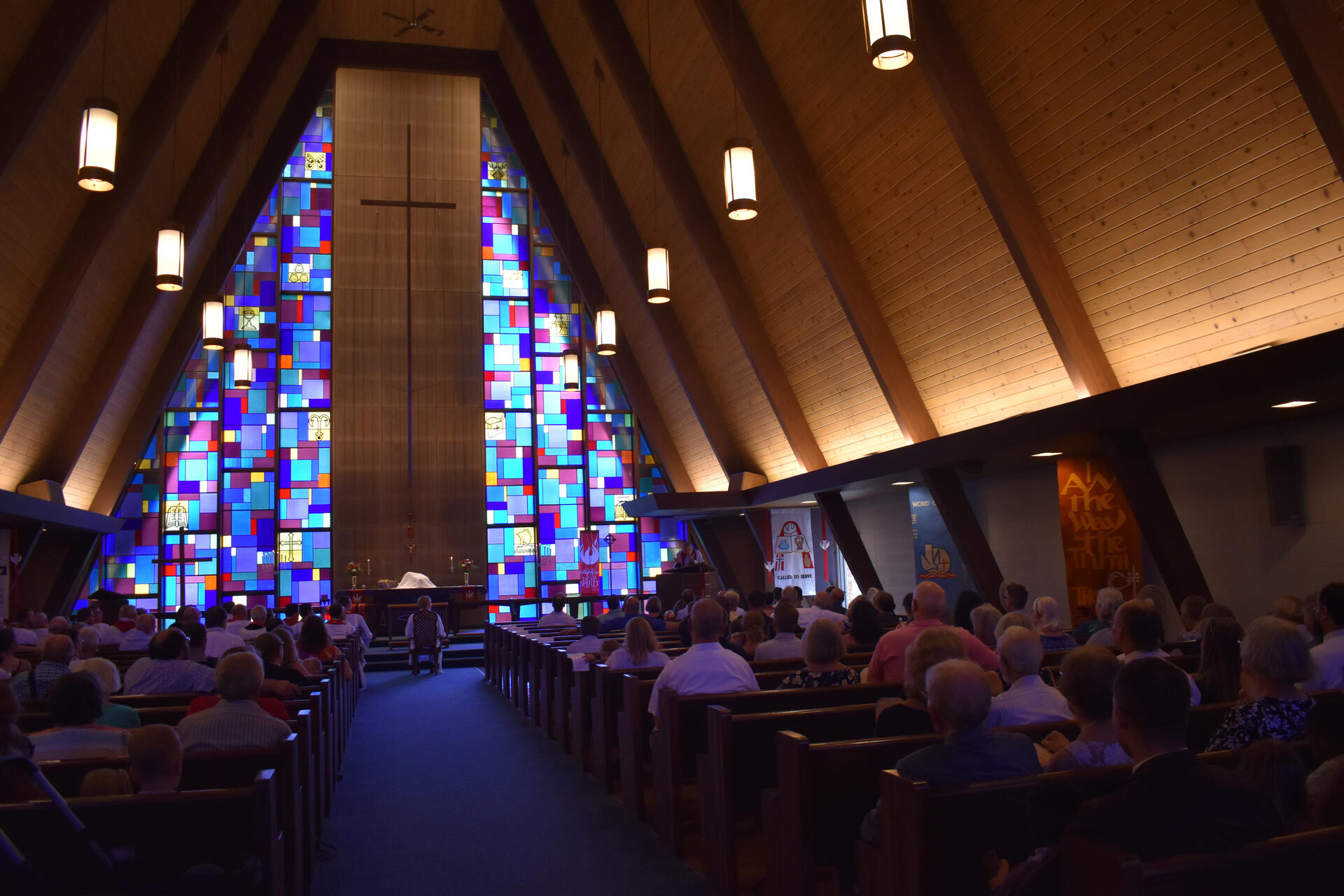 The sanctuary at Holy Cross Lutheran Church in Racine, Wisconsin during a worship service. This is from the installment of Pastor Luke Otten. He sits in a chair at the center front of the nave. The church is full, with many decorative banners, and blue and