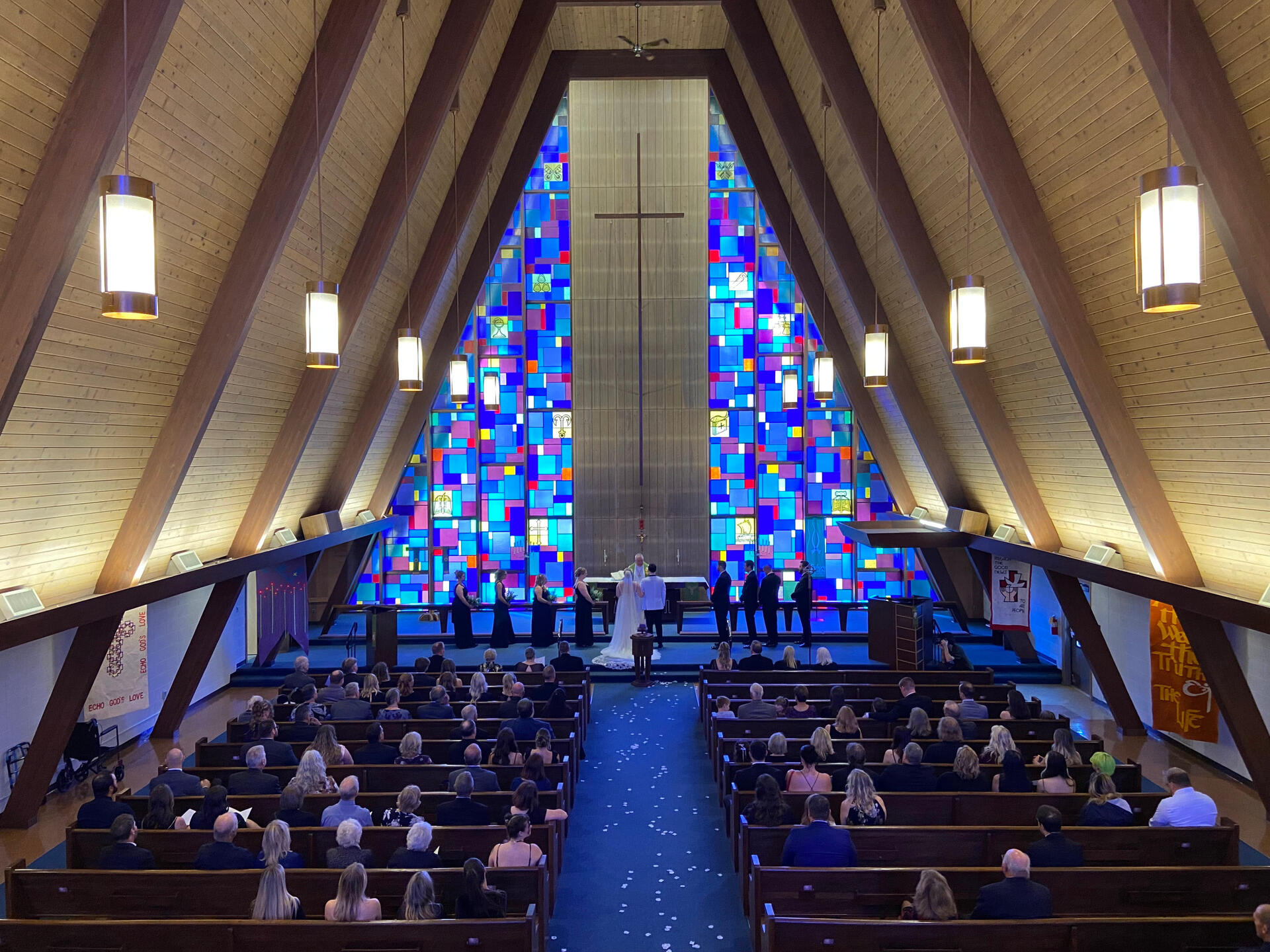 A bride and groom stand in front of the altar at Holy Cross Lutheran Church in Racine, Wisconsin, surrounded by bridesmaids and groomsmen. The blue aisle carpet is covered in flower petals, and the church is full of guests. The blue and purple stained glas