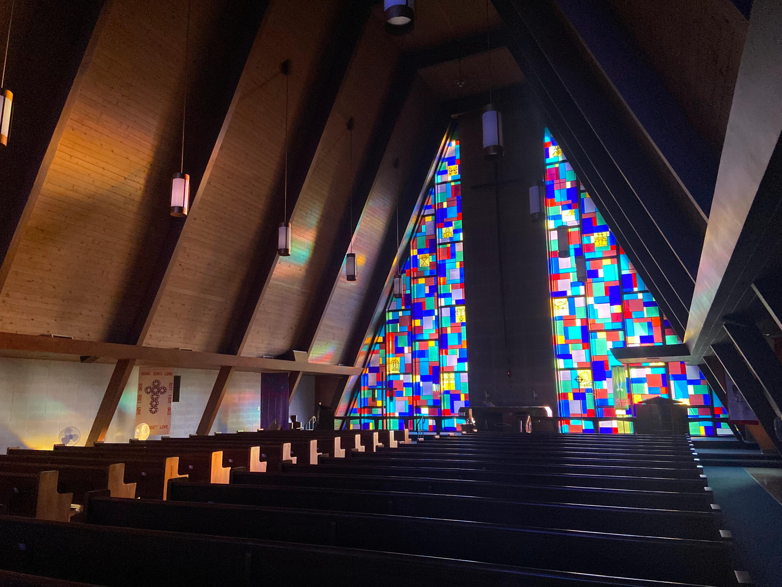 The sanctuary at Holy Cross Lutheran Church in Racine, Wisconsin. It's lit only through the striking stained glass, but with enough light to see the paneled A-frame ceiling and wooden pews.